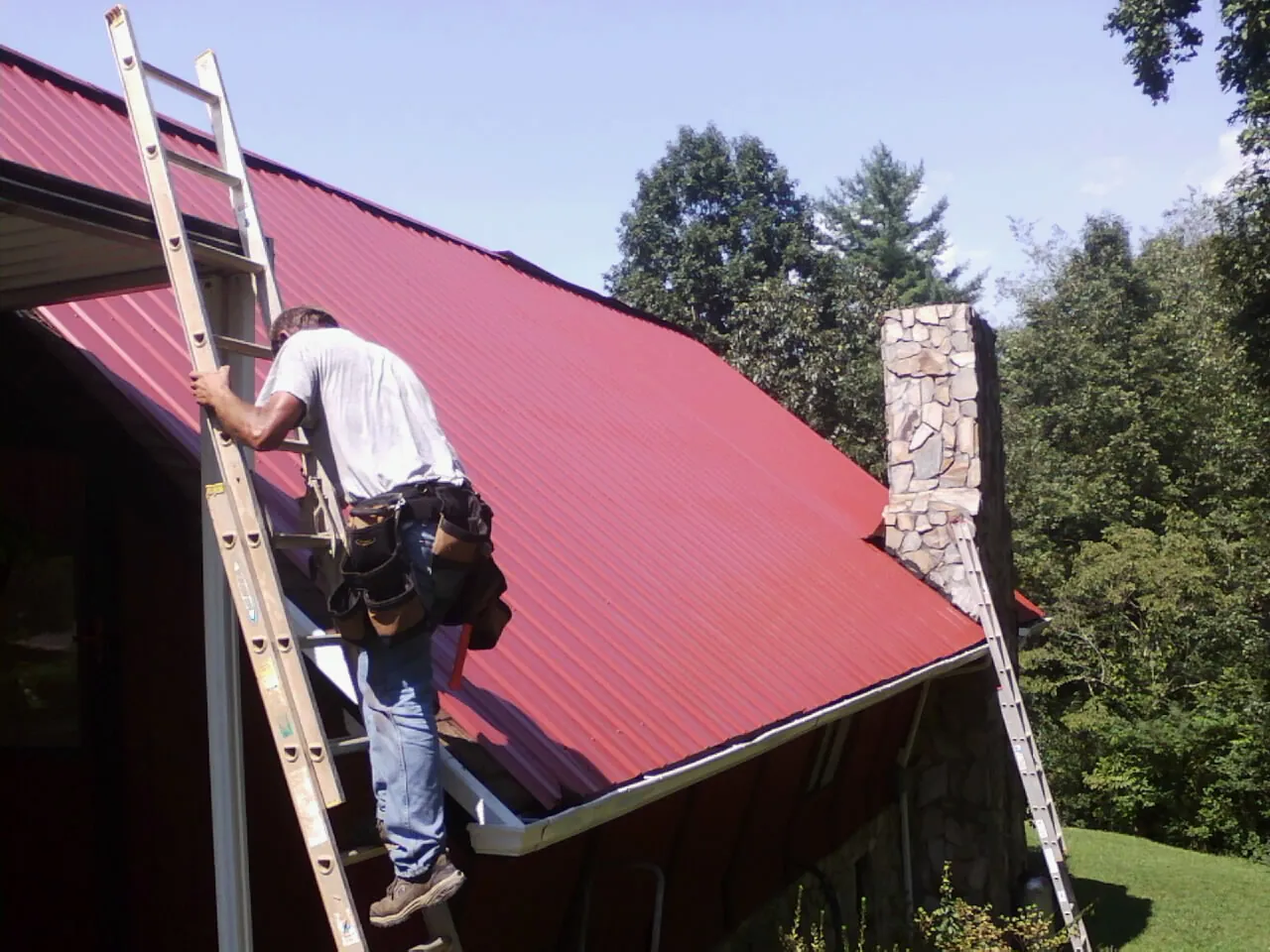 Roofer on a ladder beside a red metal roof and stone chimney — Soffit & Fascia Repair by Roofing Brothers in Zirconia